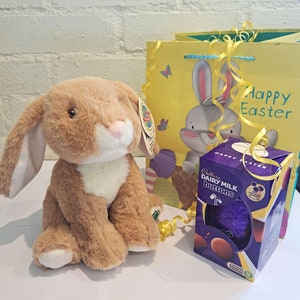 May include: A brown plush bunny rabbit toy with a white chest, sitting next to a yellow gift bag with a bunny rabbit design and the text "Happy Easter".  The gift bag is tied with a yellow ribbon.  A purple box with the text "Cadbury Dairy Milk Buttons" and a purple chocolate egg is also in the image.