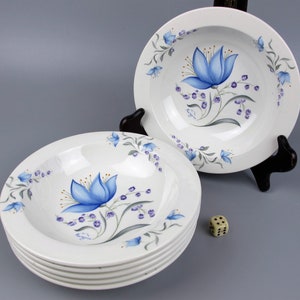 May include: A collection of white ceramic bowls, each adorned with a hand-painted blue floral motif. The bowls showcase a central blue flower and smaller floral details around the edge. The bowls are stacked, presented on a grey surface.