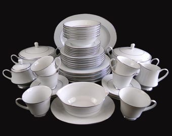 Op de afbeelding: Een compleet wit porseleinen servies met een zilveren rand. De set bevat borden, kommen, kopjes, schotels, serveerschalen en afgedekte serveerschalen. De gerechten staan op een zwarte achtergrond.