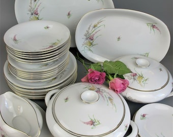 Op de afbeelding: Een set wit porselein serviesgoed met een bloemenpatroon in roze, groene en gele tinten. De set bevat borden, kommen, een serveerschaal, een sauskom en een serveerschaal met deksel. De randen van de stukken zijn versierd met een dunne gouden band.