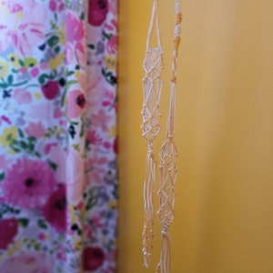 Könnte beinhalten: Zwei Makramee-Pflanzenhänger in Creme- und Brauntönen. Die Hänger sind aus geknotetem Seil gefertigt und hängen an einer Wand.