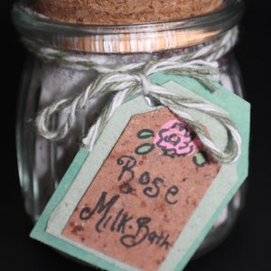 May include: A glass jar with a cork lid filled with a white powder and tied with twine and a green tag that says "Rose Milk Bath".