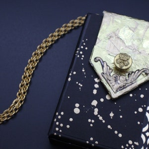 May include: A black clutch purse with a gold chain strap. The purse has a gold and white patterned design and a gold button with a floral design.