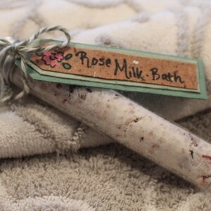May include: A glass tube filled with white bath salts and pink flower petals, tied with twine and a tag that reads "Rose Milk Bath".