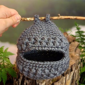 May include: A grey crocheted birdhouse hanging from a branch. The birdhouse has a round opening and is made with a textured stitch.