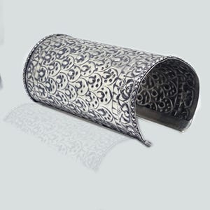 May include: Silver cuff bracelet with an intricate, repeating floral pattern. The open-ended bracelet has a curved shape and is designed to be worn on the wrist. The metal has a slightly oxidized appearance, enhancing the details of the design.