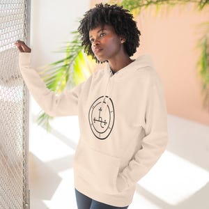 May include: Cream-colored hoodie with a black circular graphic on the front. The graphic features a cross-like symbol within a circle. The hoodie has a front pocket and a hood with a drawstring. The person is wearing blue jeans.