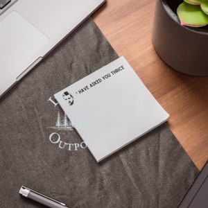 May include: White sticky note with the text "I have asked you thrice" and a black and white illustration of a man's face. The note is on a dark gray surface with a blue and white logo that says "Outpost".