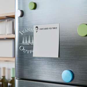 May include: A white refrigerator with a white sticker that says "I have asked you thrice" and a black and white logo that says "Ivanhoe Outpost". There are three green and one blue round magnets on the refrigerator.