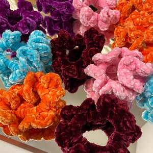 May include: Assortment of colorful scrunchies made of soft, fuzzy fabric. The scrunchies are in shades of purple, pink, blue, orange, and burgundy.