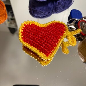 Può includere: Un cuore rosso all'uncinetto con un bordo giallo, attaccato a un portachiavi.