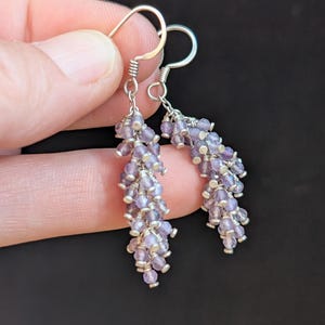 May include: A pair of dangle earrings with a cluster of small, light purple beads. The beads are strung together with silver-colored wire, and the earrings have silver-colored ear wires. The background is a solid black.
