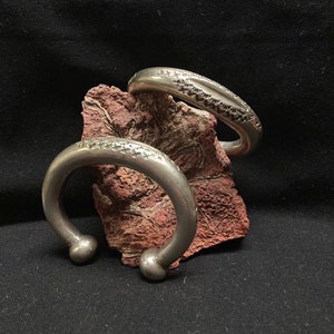 May include: Three silver bracelets with intricate designs. The bracelets are stacked on top of each other and are resting on a red rock.