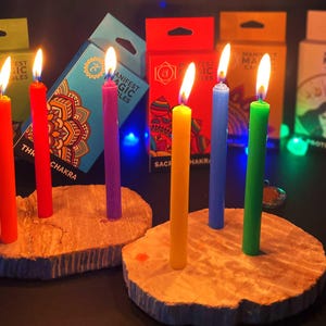 Könnte beinhalten: Brennende, farbige Kerzen auf Holzsockeln. Die Kerzen sind rot, orange, gelb, grün, blau und lila. Im Hintergrund befinden sich Schachteln mit Text und Designs, auf denen die Worte "Manifest Magic Candles" zu sehen sind.
