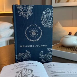 Puede incluir: Un diario de bienestar azul marino con diseños celestiales y zodiacales plateados en relieve. El diario está abierto en una página con un mandala colorido y indicaciones de registro diario.