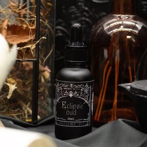 May include: A matte black glass dropper bottle labelled Eclipse Oud 30ml perfume oil. The label features white ornate decorative borders and text. The bottle is positioned among dark objects including a glass lantern and a brown glass bottle.