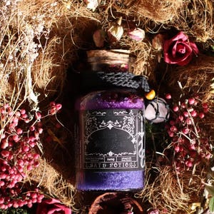 Op de afbeelding: Een glazen pot gevuld met paars badzout, afgesloten met een kurk en een zwart koord. Het zwarte etiket bevat de witte tekst Love Bath Spell en 240g. De pot is omringd door gedroogde plantenvezels, roze bessen en rode rozen.