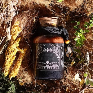 Puede incluir: Un frasco de vidrio con tapón de corcho que contiene un producto de baño en capas. La etiqueta negra presenta el texto Success Bath Spell y 240g. Un cordón de ganchillo negro con una cuenta de madera envuelve el cuello del frasco, que contiene un producto naranja y negro.