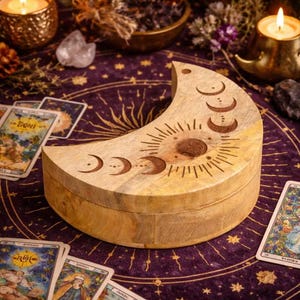 May include: A light brown, crescent moon-shaped wooden box, etched with sun and moon phase designs. The box is on a purple cloth with gold star patterns. Tarot cards and candles surround the box, creating a mystical theme.