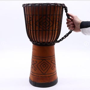 Może przedstawiać: Brązowy bęben djembe z dekoracyjnym geometrycznym wzorem. Bęben ma drewniany korpus i ciemnobrązowy uchwyt z liny. Korpus bębna jest ozdobiony czarnymi geometrycznymi wzorami. Jest to instrument perkusyjny.