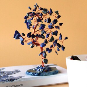 May include: A decorative tree sculpture with a copper wire trunk and branches, adorned with blue gemstone leaves. The tree sits on a blue stone base, placed on a book with the words "ANCIENT WISDOM". The background is a warm, neutral color.