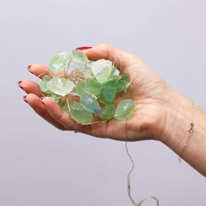 Könnte beinhalten: Eine Handvoll roher, grüner Fluoritkristalle auf einem feinen Draht mit kleinen Lichtern. Die Kristalle variieren in Grüntönen und werden von den winzigen Lichtern beleuchtet. Ein goldenes Armband ist am Handgelenk zu sehen.