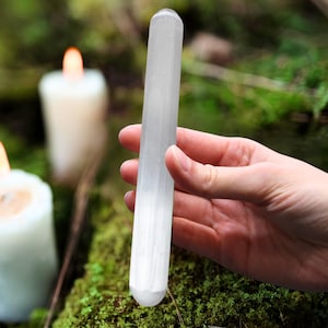 Op de afbeelding: Een heldere, langwerpige selenietstaaf wordt in een hand gehouden tegen een vage achtergrond van groen mos en brandende kaarsen. De staaf is cilindrisch met een afgeronde punt en een getextureerd, kristallijn uiterlijk. De scène suggereert natuur en spirituele praktijken.