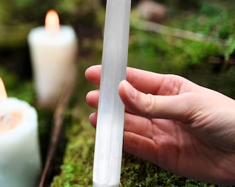 Op de afbeelding: Een heldere, langwerpige selenietstaaf wordt in een hand gehouden tegen een vage achtergrond van groen mos en brandende kaarsen. De staaf is cilindrisch met een afgeronde punt en een getextureerd, kristallijn uiterlijk. De scène suggereert natuur en spirituele praktijken.