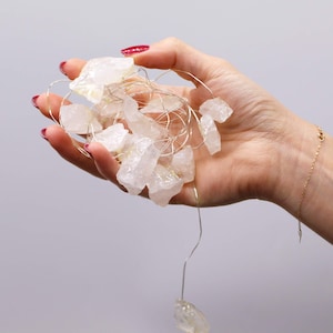 Pode incluir: Uma mão segurando uma corda de luzes com cristais de quartzo transparentes e brutos. Os cristais têm formato irregular e variam em tamanho. O fio é prateado. O fundo é cinza claro liso.