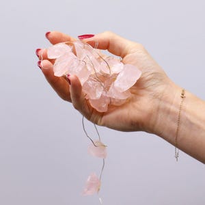 Può includere: Una mano tiene in mano una stringa di luci di cristallo di quarzo rosa. I cristalli sono di forma irregolare e di dimensioni variabili. Un delicato braccialetto d'oro è visibile sul polso. Lo sfondo è di un colore neutro.