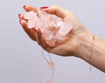 Op de afbeelding: Een hand houdt een slinger van roze rozenkwarts kristallen lichten vast. De kristallen zijn onregelmatig van vorm en variëren in grootte. Een delicate gouden armband is zichtbaar om de pols. De achtergrond is een zachte, neutrale kleur.