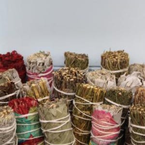 Può includere: Una collezione di vari fasci di salvia, ben avvolti con spago e tessuto colorato. I fasci mostrano una gamma di colori, tra cui verde, rosso e bianco, suggerendo diversi tipi di erbe e botaniche. L'immagine è ripresa da vicino.