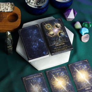 Può includere: Un mazzo di "Celestial Grace Cards" con un design a notte stellata, con pianeti e il testo "Celestial Grace Cards". Le carte sono esposte con cristalli, candele e altri oggetti spirituali su una superficie verde.