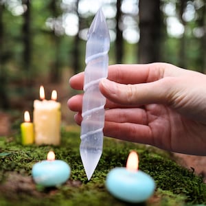 Puede incluir: Una varita de cristal de selenita transparente con un diseño en espiral se sostiene en una mano. La varita tiene una punta en un extremo. Velas encendidas de varios tamaños y colores están en el fondo, lo que sugiere un tema espiritual o decorativo.