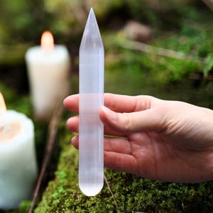 Puede incluir: Una varita de cristal de selenita transparente, de unos 15 cm de largo, se sostiene en una mano. La varita tiene un extremo puntiagudo y una forma cilíndrica. Dos velas encendidas están en el fondo, con musgo verde y follaje.