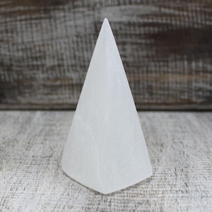 Può includere: Un cristallo di selenite bianco intagliato in una forma a cono liscia e appuntita. Il minerale presenta una consistenza fibrosa e traslucida con sottili variazioni naturali sulla superficie opaca, eretto con una base circolare e un apice.