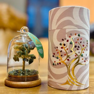 May include: A decorative glass cloche with a small tree sculpture made of green stones and gold wire, set on a wooden base. Beside it is a cylindrical container with a tree design and colorful stone accents.
