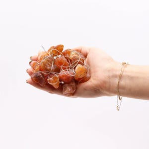 Op de afbeelding: Een handvol ruwe, oranje-bruine carneool edelstenen, verlicht door kleine witte lampjes. Een delicate gouden armband met bedels siert de pols. De edelstenen worden tegen een witte achtergrond gehouden.