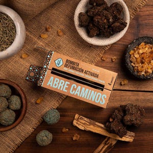 Puede incluir: Una caja de cartón rectangular etiquetada como "BOMBITA DEFUMACIÓN ACTIVADA" y "ABRE CAMINOS" que contiene cuatro esferas de incienso, rodeada de cuencos con hierbas secas, trozos de resina y palos sobre arpillera.