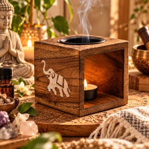 May include: A square wooden oil burner featuring a carved elephant inlay on the side. The burner has a circular black metal dish on top for essential oils and an open front compartment containing a lit tea light candle emitting thin smoke.