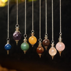 Op de afbeelding: Een verzameling edelsteen slingerpendels die aan zilveren kettingen hangen. Elke slinger heeft een edelsteen van een andere kleur, waaronder blauw, paars, groen, oranje, bruin, helder en roze. De pendels hebben een druppelvormige metalen punt.