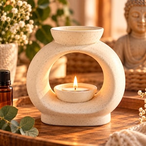 Puede incluir: Un quemador de aceite de cerámica color crema con acabado moteado y marco circular. El quemador cuenta con un cuenco superior para aceite y una abertura central que sostiene una pequeña vela de té encendida, creando un brillo cálido.