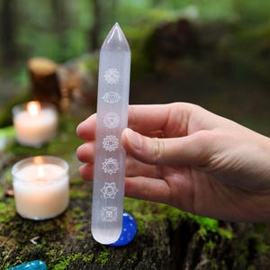 Könnte beinhalten: Ein klarer, länglicher Selenit-Kristallstab mit geätzten Chakra-Symbolen. Der Stab wird in einer Hand gehalten, mit einem verschwommenen Hintergrund aus Kerzen und Grün. Der Kristall ist etwa 15 cm lang.