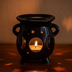 Peut inclure: Un photophore noir en céramique en forme de chaudron avec une bougie allumée à l'intérieur. Le chaudron a deux poignées et est décoré de découpes en forme de croissant de lune et d'étoiles. Le photophore repose sur trois petits pieds.