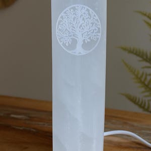 Puede incluir: Lámpara rectangular blanca de selenita con un diseño de árbol de la vida tallado. La lámpara está iluminada y tiene un cable de alimentación. La lámpara está sobre una superficie de madera. La lámpara mide unos 20 cm de altura.