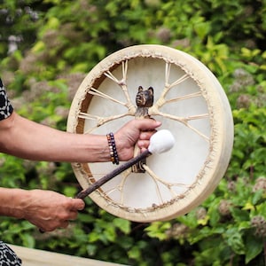 May include: A round, cream-colored drum with a wooden handle carved with a bear figure. The drum is held with one hand, and a mallet with a white head is held in the other hand. The drum is decorated with natural-colored twine.