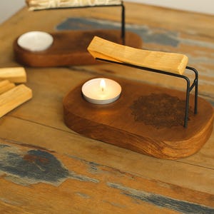 May include: Wooden candle holders with black metal supports, holding a lit tea light and a piece of palo santo. The holders are on a wooden surface, with a mandala design etched into the wood.