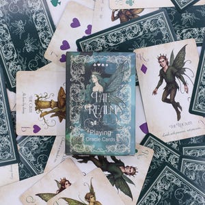 Pode incluir: Um baralho de "Fae Realm Playing Oracle Cards" com uma caixa verde-azulado e prateada com uma ilustração de fada. As cartas estão espalhadas ao redor da caixa, com várias ilustrações e texto temáticos de fadas.