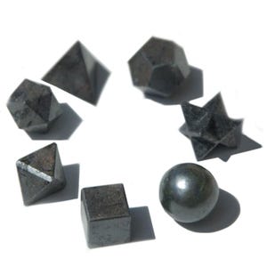 Puede incluir: Un surtido de formas geométricas de color gris oscuro, que incluyen una pirámide, un cubo, una esfera y varios poliedros. Los objetos están dispuestos sobre un fondo blanco, proyectando sombras. Las formas varían en tamaño y tienen un aspecto metálico.