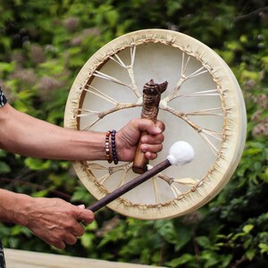 Op de afbeelding: Een ronde, crèmekleurige trommel met een houten frame en een gesneden handvat. De trommel wordt door een persoon vastgehouden, met een hamer in de andere hand. De achtergrond is wazig groen gebladerte.